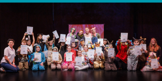 Eine Gruppe Kinder sitzt in bunten Kostümen auf einer Bühne, lächeln in die Kamera und haben Urkunden in der Hand.