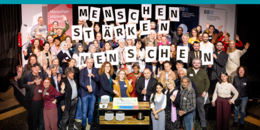 eine größere Gruppe an Menschen steht in mehreren Reihen hintereinander, alle blicken in die Kamera, viele von ihnen halten zusammen Schilder mit einzelnen Buchstaben hoch, die zusammen den Text "Menschen stärken Menschen" ergeben