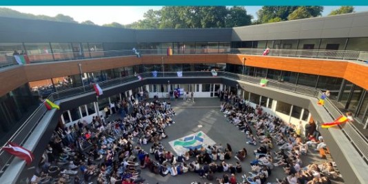 Auf einem Schulhof sind viele Menschen versammelt. Das Gebäude ist mit Flaggen verschiedener Länder geschmückt.
