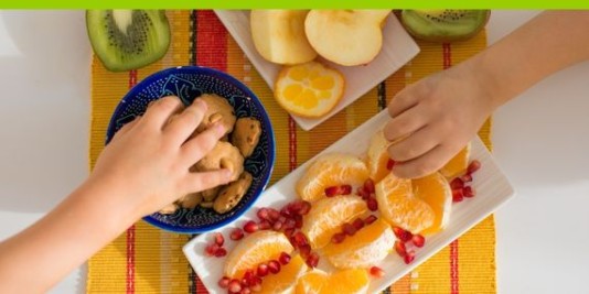 Zwei Kinderhände greifen nach Tellern, die mit Obst und Keksen gefüllt sind. 