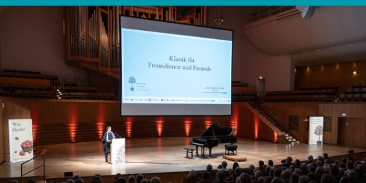 Auf einer Bühne steht eine Person und hält eine Rede, neben ihr steht ein Flügel. Über der Bühne ist eine große Leinwand, auf der "Klassik für Freundinnen und Freunde" steht.