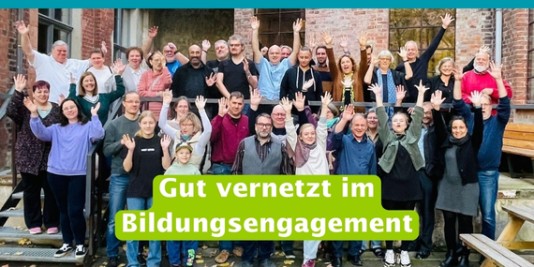 Eine Gruppe von Menschen steht vor einem Gebäude, lächelt in die Kamera und hebt die Arme in die Luft. Davor die Aufschrift: Gut vernetzt im Bildungsengagement
