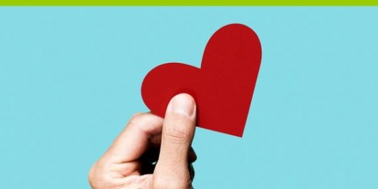 Eine Hand hält ein rotes Herz aus Papier zwischen Daumen und Zeigefinger.