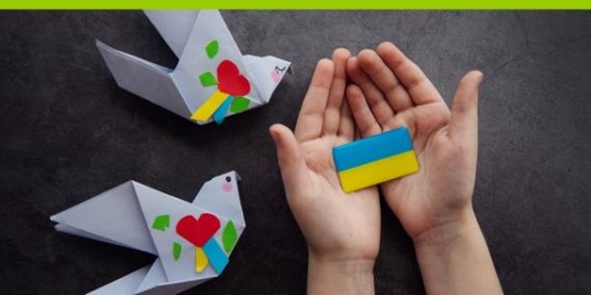 Kinderhände halten eine kleine Ukraineflagge in blau und gelb. Zwei aus Papier gefaltete und mit Herzen beklebte Tauben liegen daneben.