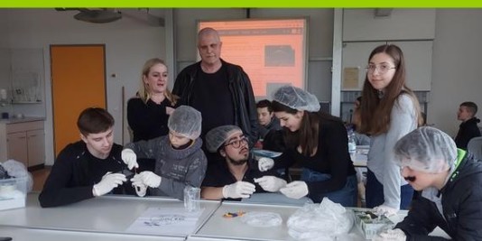 Jugendliche und Kinder mit Haarnetzen und weißen Handschuhen arbeiten an einem Tisch in einem Klassenraum. Hinter ihnen stehen zwei Erwachsene.