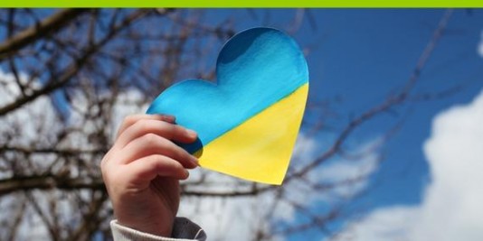 Eine Kinderhand hält ein Papierherz in blau und geld, den Farben der ukrainischen Flagge, in die Luft