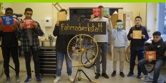 Jungen stehen in einer Reihe und halten bunte Karten in den Händen. In ihrer Mitte steht ein Schild mit der Aufschrift Fahrradwerkstatt.