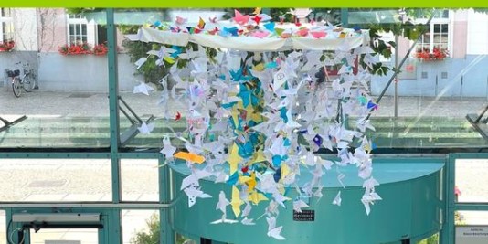 Hunderte Origami-Tauben hängen an einem großen Mobile angeordnet in der Luft vor einer verglasten Wand.