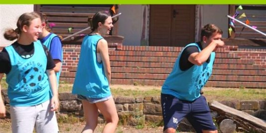 Vier Jugendliche stehen auf einem Hof in der Sonne. Sie tragen türkisfarbene Trikots mit einem Fußball drauf. Sie lachen.