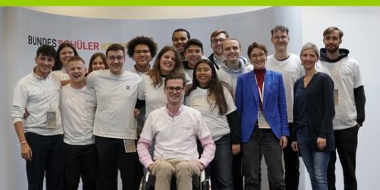 Eine Gruppe junger Menschen steht vor einer weißen Wand mit der Aufschrift Bundesschülerkonferenz in einer Gruppe und lächelt in die Kamera.