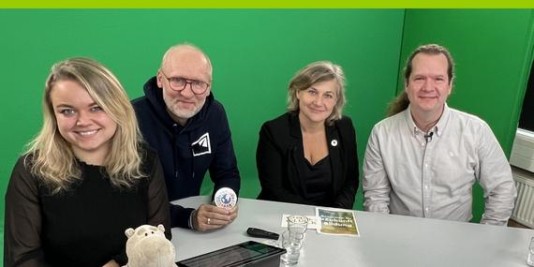 Vier Personen sitzen an einem Tisch vor einer grünen Wand und lächeln in die Kamera. Eine Person ist Katja Hintze.