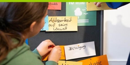Ein Mädchen pinnt einen Zettel an eine Wand. Andere Zettel sind zu lesen: Beteiligung, Achtsamkeit auf seine Umwelt