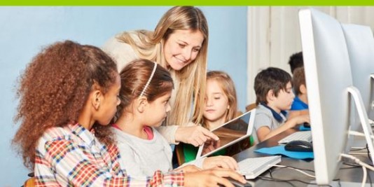 Eine Lehrerin zeigt Kindern, die vor Computerbildschirmen stzen, etwas auf einem Tablet