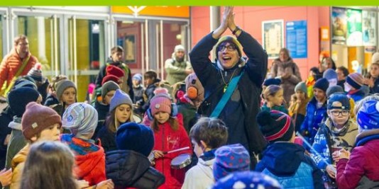 Viele Kinder in Winterkleidung stehen mit Schlaginstrumenten zusammen. Ein Erwachsener in der Mitte klascht über seinem Kopf in die Hände.
