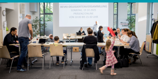 mehrere Menschen sitzen auf Stühlen an Tischen vor einem großen Screen, auf dem eine Präsentation der bevki läuft, ein Kind läuft hinter ihnen vorbei