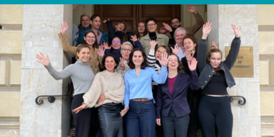 eine größere Gruppe an Menschen steht in mehreren Reihen hintereinander, alle lächeln in die Kamera und heben die Arme hoch