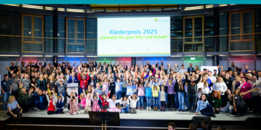Eine große Gruppe Menschen steht auf einer Bühne und hebt glücklich die Arme. Über ihnen ist eine Leinwand mit der Aufschrift „Förderpreis 2025 Verein(t) für gute Kita und Schule“