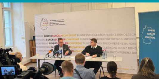 Quentin Gärtner, Generalsekretär der Bundesschülerkonferenz, und Prof. Dr. Michael Hüther, Direktor des Instituts der deutschen Wirtschaft (IW), sitzen umringt von Mikrofonen an einem Tisch. Ihnen gegenüber sitzen viele Menschen und viele Kameras. Hinter ihnen ist ein Banner der Bundesschülerkonferenz.
