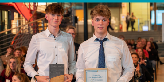 zwei junge Menschen in weißen Hemden stehen vor sitzendem Publikum, sie halten einen Pokal und eine Urkunde in Händen