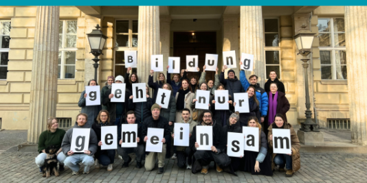 Eine Gruppe Menschen steht vor einem Gebäude. Sie halten Buchstaben in der Hand, die den Text „Bildung geht nur gemeinsam" bilden