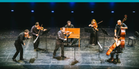 Sieben Musiker*innen musizieren gemeinsam auf einer Bühne. Sie sind alle schwarz angezogen. Die Instrumente sind Geigen, Bratsche, Cello, Kontrabass und Klavier.