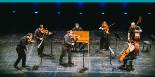 Sieben Musiker*innen musizieren gemeinsam auf einer Bühne. Sie sind alle schwarz angezogen. Die Instrumente sind Geigen, Bratsche, Cello, Kontrabass und Klavier.