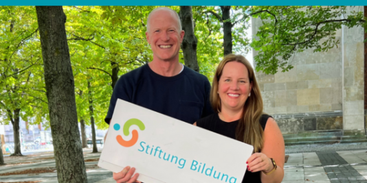 Zwei Menschen stehen bei gutem Wetter draußen vor Bäumen und lächeln in die Kamera. Sie halten ein Schild in den Händen, auf dem das Logo der Stiftung Bildung zu sehen ist sowie deren Schriftzug.