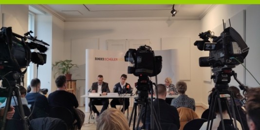 Vier Menschen sitzen an einem Tisch vor einer weißen Wand. Vor ihnen stehen Kameras und Menschen sitzen ihnen zugewandt in Stuhlreihen.