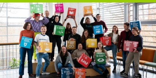 Eine Gruppe junger Menschen steht vor einer Fensterwand und lächelt in die Kamera.