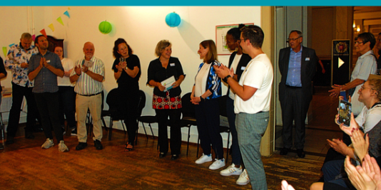 mehrere Menschen stehend und sitzend applaudieren, im Hintergrund bunte Deko