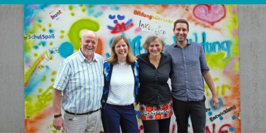 4 Menschen stehen nebeneinander, Arm in Arm und lächeln nach vorne in die Kamera