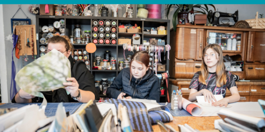 drei junge Menschen sitzen an einem Tisch in einer Bastelwerkstatt, vor ihnen auf dem Tisch und hinter ihnen im Regal viel Material