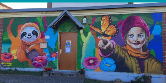 Eine bunt bemalte Mauer vor einem Jugendclub mit einem Faultier, Blumen, einem Schmetterlink und einem Mädchen mit roter Mütze.