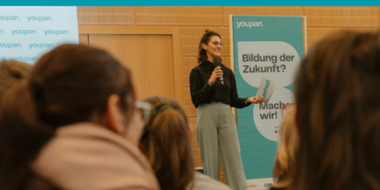 Eine junge Frau steht vor einer Gruppe Menschen und redet ins Mikrofon. Hinter ihr ist ein blaues Banner mit der Aufschrift: youpan. Bildung der Zukunft? Machen wir!