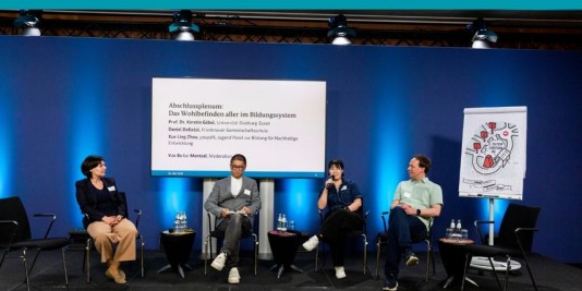 Auf einer Bühne sitzen vier Menschen auf Stühlen in einer Gesprächsrunde, im Hintergrund eine Bildschirmpräsentation und ein Flipchart mit einer Zeichnung.