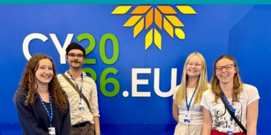 Vier junge Menschen stehen vor einer blauen Wand mit dem folgenden Schriftzug: CY2026.EU