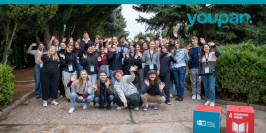 eine Gruppe aus circa 30 jungen Menschen steht eng zusammen und winkt in die Kamera, im Hintergrund Bäume, rechts oben das Logo vom youpan